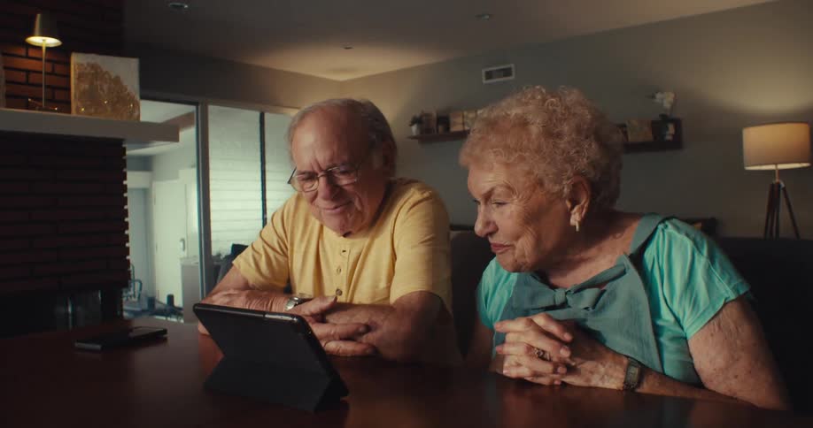 An elderly couple sit at a table in a warmly lit living room focused on a tablet device together