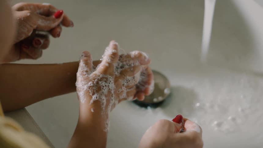 An adult and a child are washing hands together under running water in a sink with soap lather on their hands