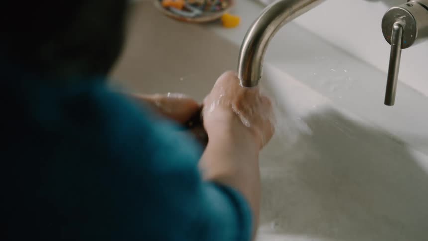 A mother stands behind her daughter and helps her wash her hands using the bathtub faucet