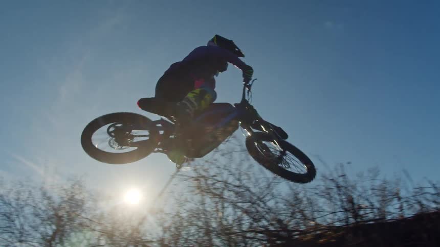 A dirtbiker jumps through the air against a clear blue sky with sunlight behind him