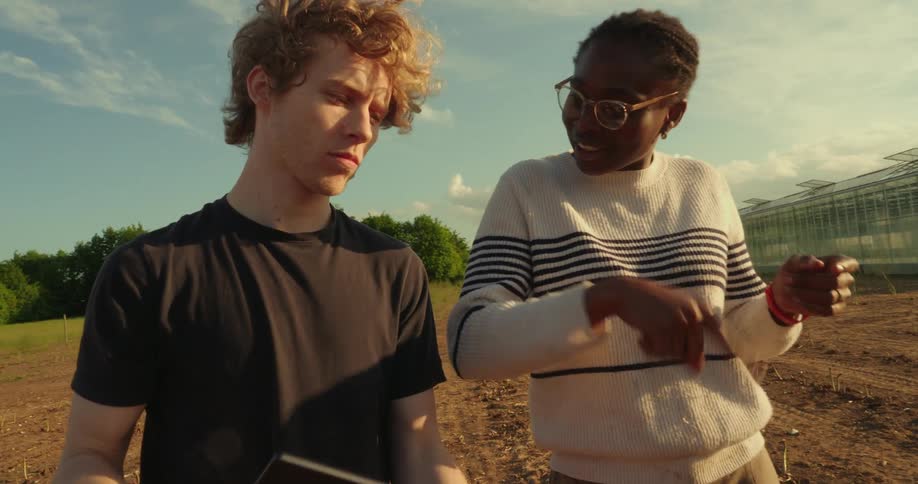 Two people are standing in a farm field discussing something while looking at a laptop