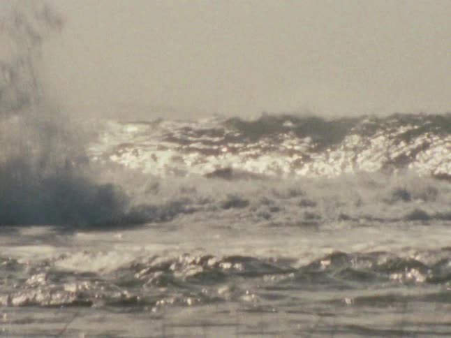 Powerful ocean waves crash against rocks under a hazy sky
