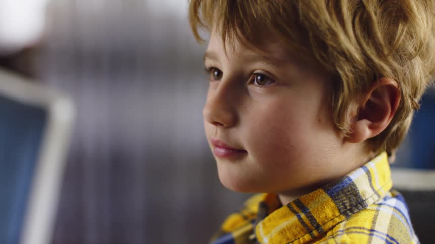 A child with blonde hair in a yellow plaid shirt sits indoors laughing as a hand playfully intercepts his own midmotion while he tries to eat a booger