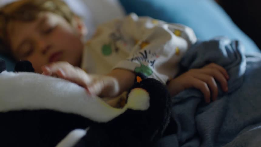 A child sleeping in bed under a blanket picks up a teddy bear beside him and goes back to sleep