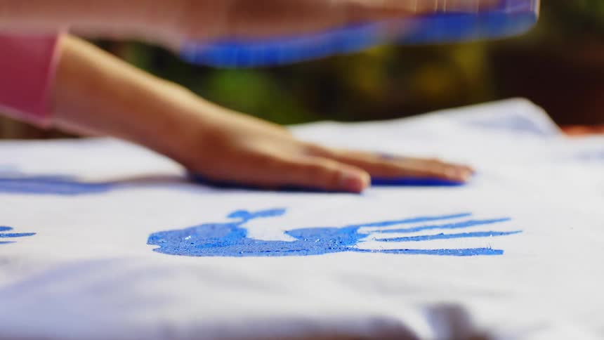 Close up of a persons hands as they place blue palm prints on a piece of clothing on a surface outdoors during daytime