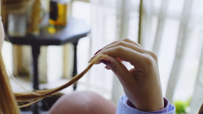Woman sitting near a curtained window plays with a lock of her hair