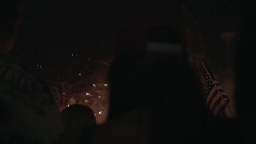 A crowd watches fireworks with an american flag waving in the background