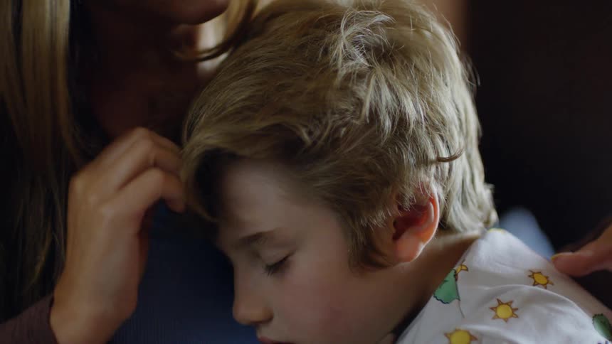 A mother gently carries her sleeping child dressed in pajamas softly rubbing his back and stroking his hair as he rests peacefully in her arms