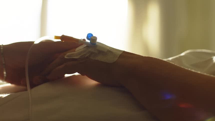 A person holds the hand of a hospital patient lying in bed with a saline drip attached