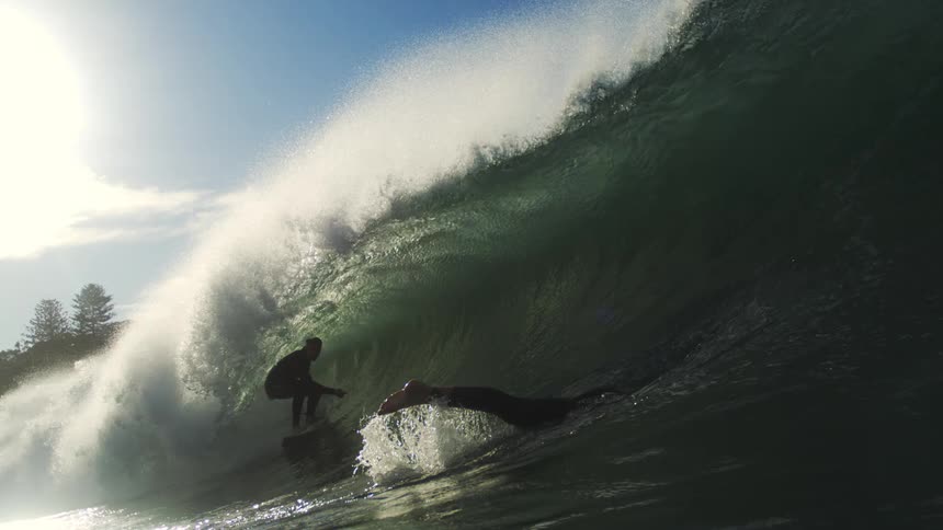Surfer rides a massive wave at sunset as another floats nearby in the curl