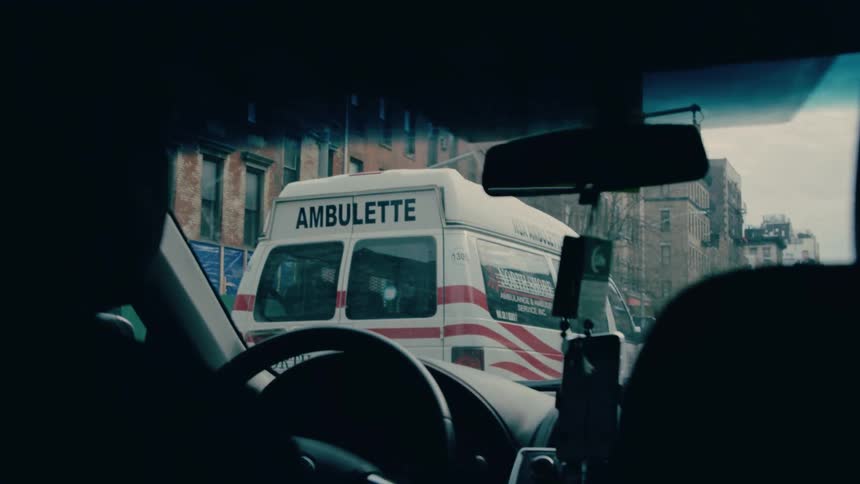 Close up of a man driving a vehicle on a city street behind an ambulance van during daytime