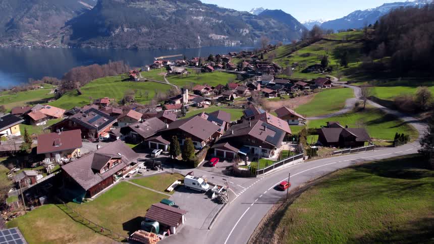 A tranquil village sits beside a reflective lake framed by rolling hills and distant snowcapped mountains under a clear sky