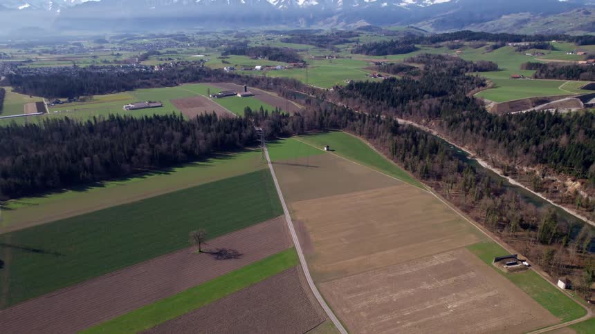 A winding river cuts through a patchwork of farmland and forest with snowcapped mountains rising in the distance