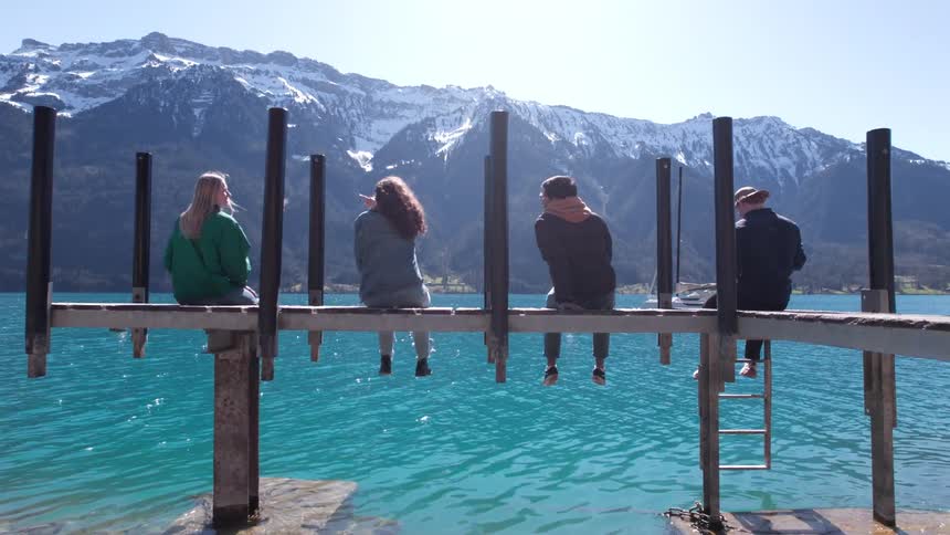 Group of four people sitting on a wooden deck by a lake with mountains in the background