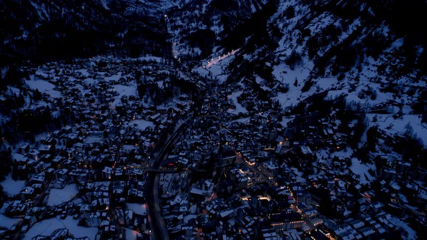 Snowcovered mountain village at dusk with warm lights glowing between forested slopes