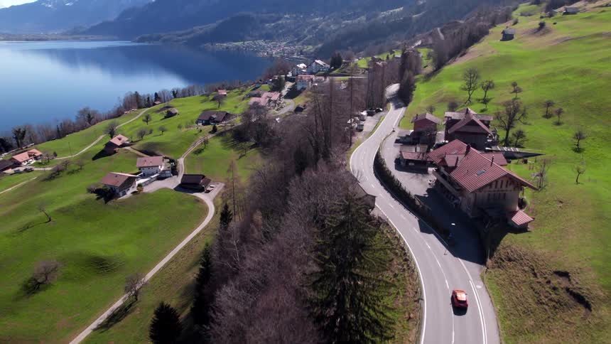 A red car drives past a small town surrounded by green vegetation at midday