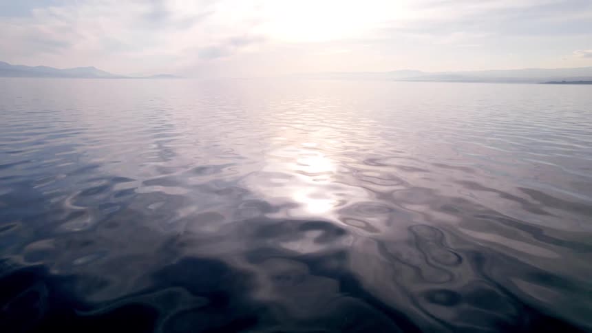 A lowangle shot glides over the calm surface of a large body of water where the bright sun creates a shimmering reflection on the gentle ripples