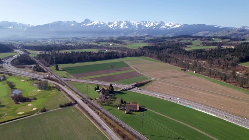 A highway cuts through farmland and scattered homes with snowcapped mountains rising in the distance under a clear sky