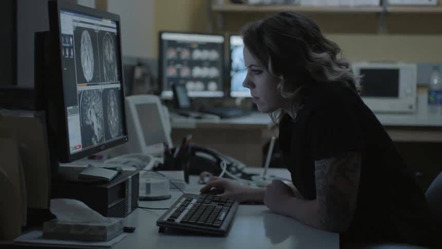 A woman sits at a desk looking at scanned images of a brain