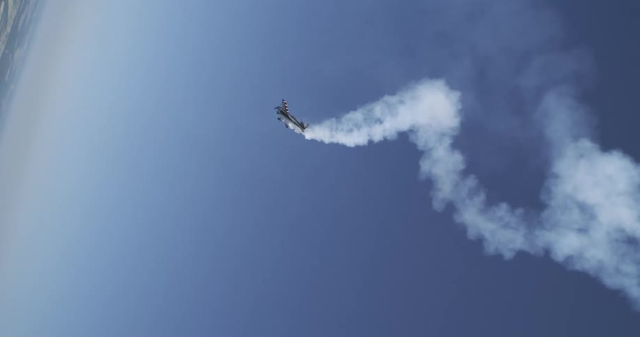 An airplane performs an aerobatic maneuver with a smoke trail curling behind it against a clear blue sky