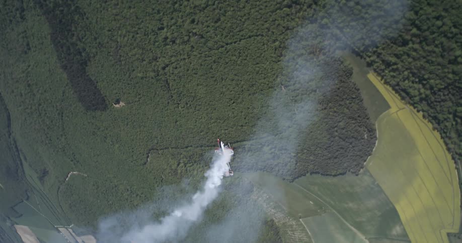 A stunt plane releases a thick smoke trail while flying over a dense forested landscape