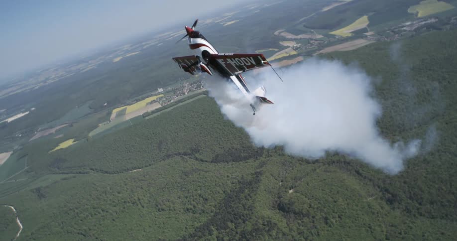 An xtremeair sbach 300 airplane soars through the air trailing smoke as it flies over lush green vegetation