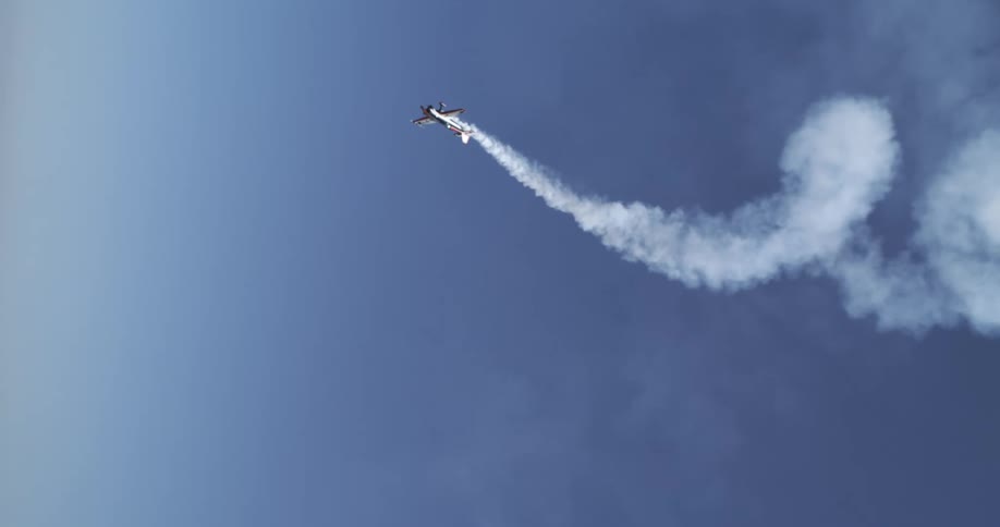 An airplane performs an aerobatic maneuver with a smoke trail curling behind it against a clear blue sky