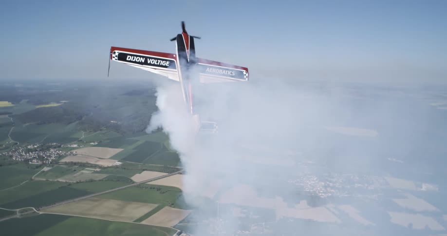 A stunt plane climbs vertically through the sky trailing thick smoke over farmlands far below