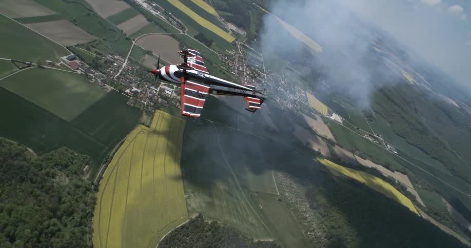 A stunt plane twists through the sky trailing smoke over patchwork farmland below