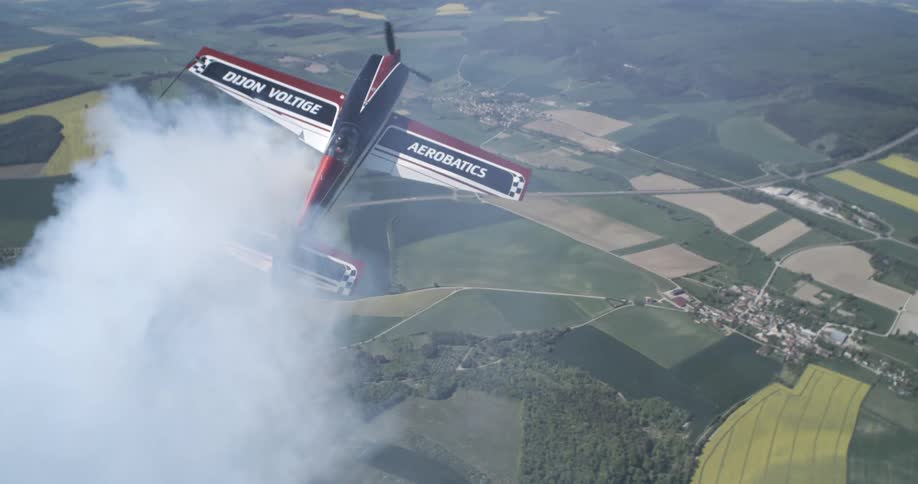 An aerobatic plane is performing a maneuver above a patchwork of fields and a small town