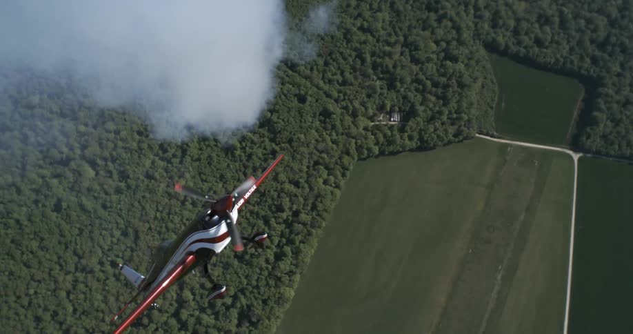 A stunt plane performs aerobatics over a dense forest landscape and farmland below