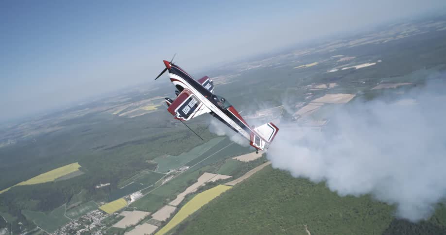 A black and white single engine plane flying over several fields of vegetation and releasing white smoke from behind