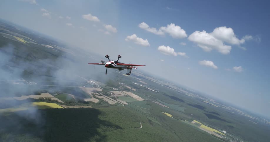 A small aerobatic airplane flies over a vast rural landscape trailing smoke