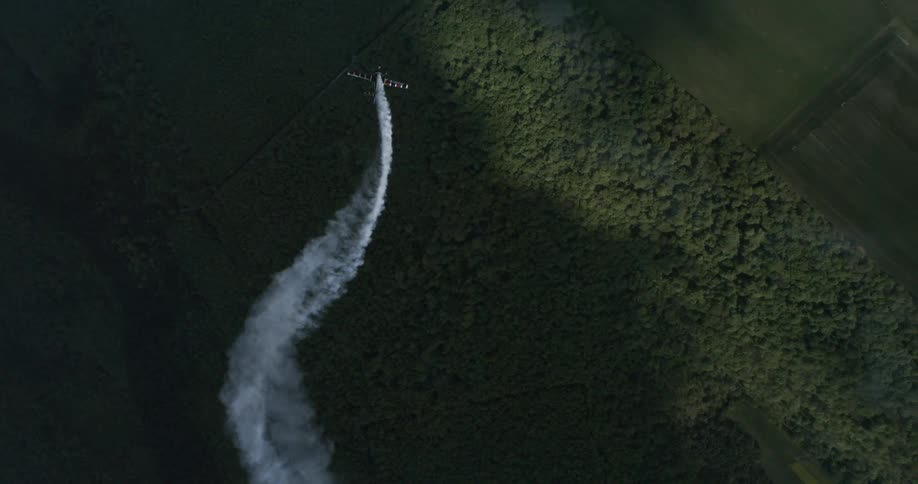An airplane trails white smoke as it flies over a dense forest captured from directly above