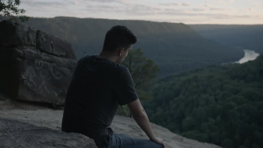 A man sits on the ground near a ledge looking out over a natural view