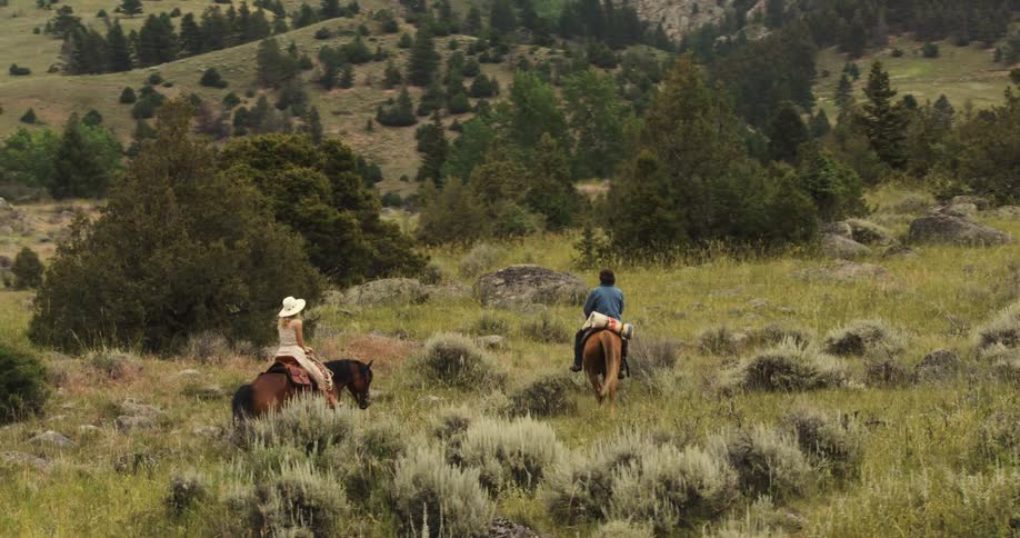 Two people ride horses through a grassy rocky meadow surrounded by hills and trees