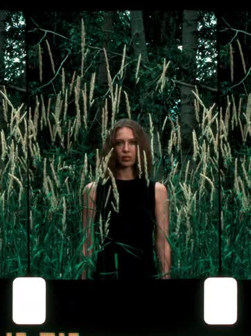 A woman stands alone in tall grass surrounded by dense forest facing the camera