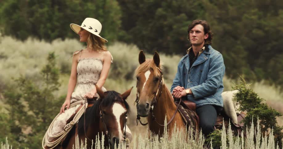 A young blonde woman in a dress and hat sits on the back of a horse and talks to a young brunet man in a jean jacket sitting on a horse beside her surrounded by wilderness with mountains and a gray sky in the background during the day
