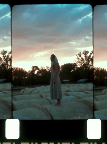 A woman standing atop a stack of straw bales in an open field under a cloudy sky