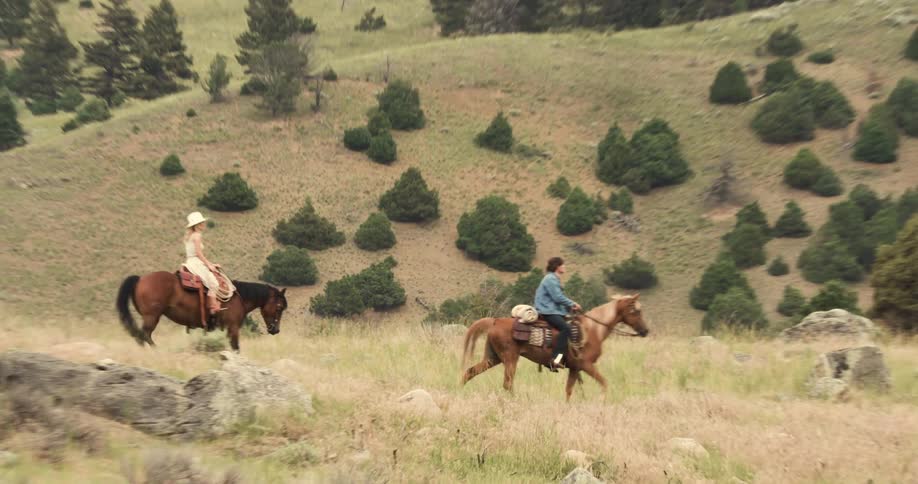 A man and a woman are riding horses in a grassy field with a hill in the background full of trees
