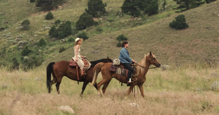 Man and woman ride horses down a hill with mountains looming behind them
