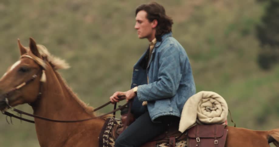 A man and a woman are riding horses in an outdoor environment and in the background there are hills with green grass and some trees