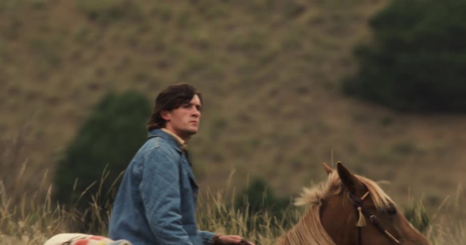 Two horseback riders cross a grassy hillside with scattered trees and rocks