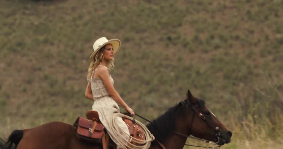 A couple ride horses along a path through a hilly semiarid landscape dotted with sparse trees and shrubs