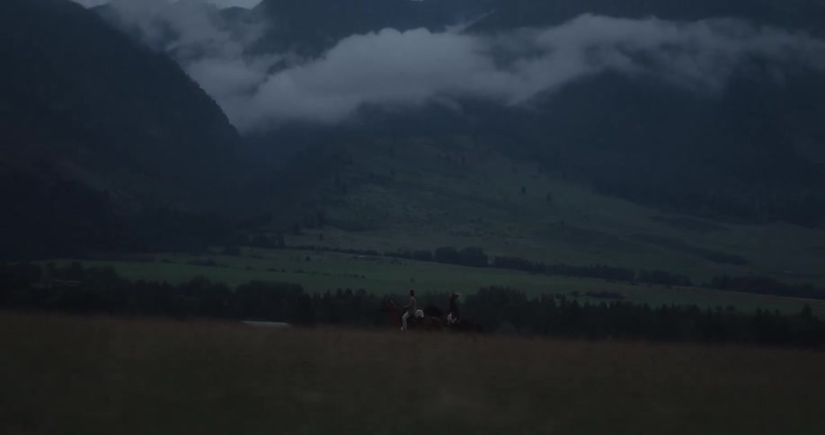 Two people ride horses across a quiet grassland at dusk with mountains in the background