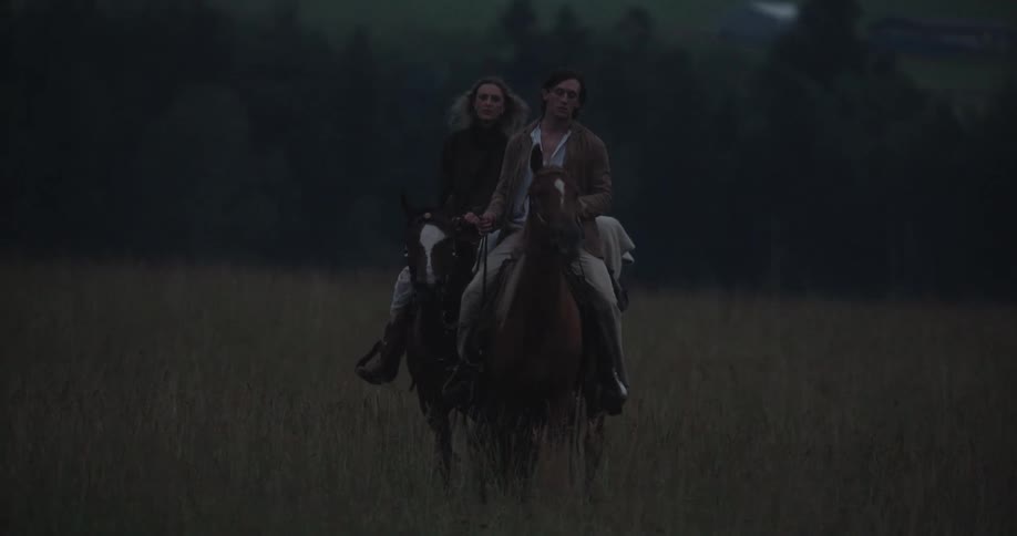 Two people ride horses across a grassy field under a twilight sky
