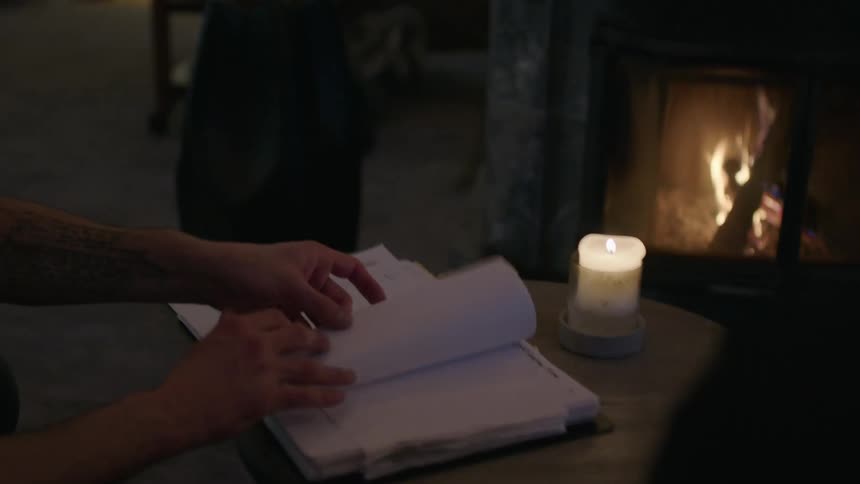 A person flips through a notebook beside a lit candle near a cozy fireplace