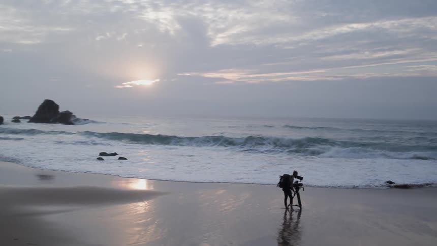 A photographer is standing on a beach with a tripod at dusk