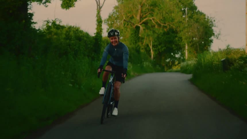 A smiling cyclist rides toward the camera along a quiet rural road surrounded by lush greenery