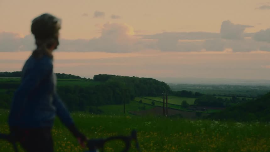 A cyclist stands still in a grassy field at sunset looking out over a distant landscape